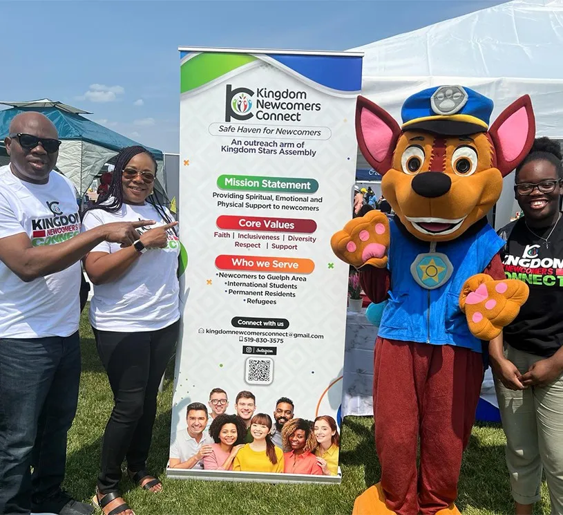 Kingdom Newcomers Connect members and a dog mascot beside their banner at a Kingdom Stars Assembly outreach, Guelph