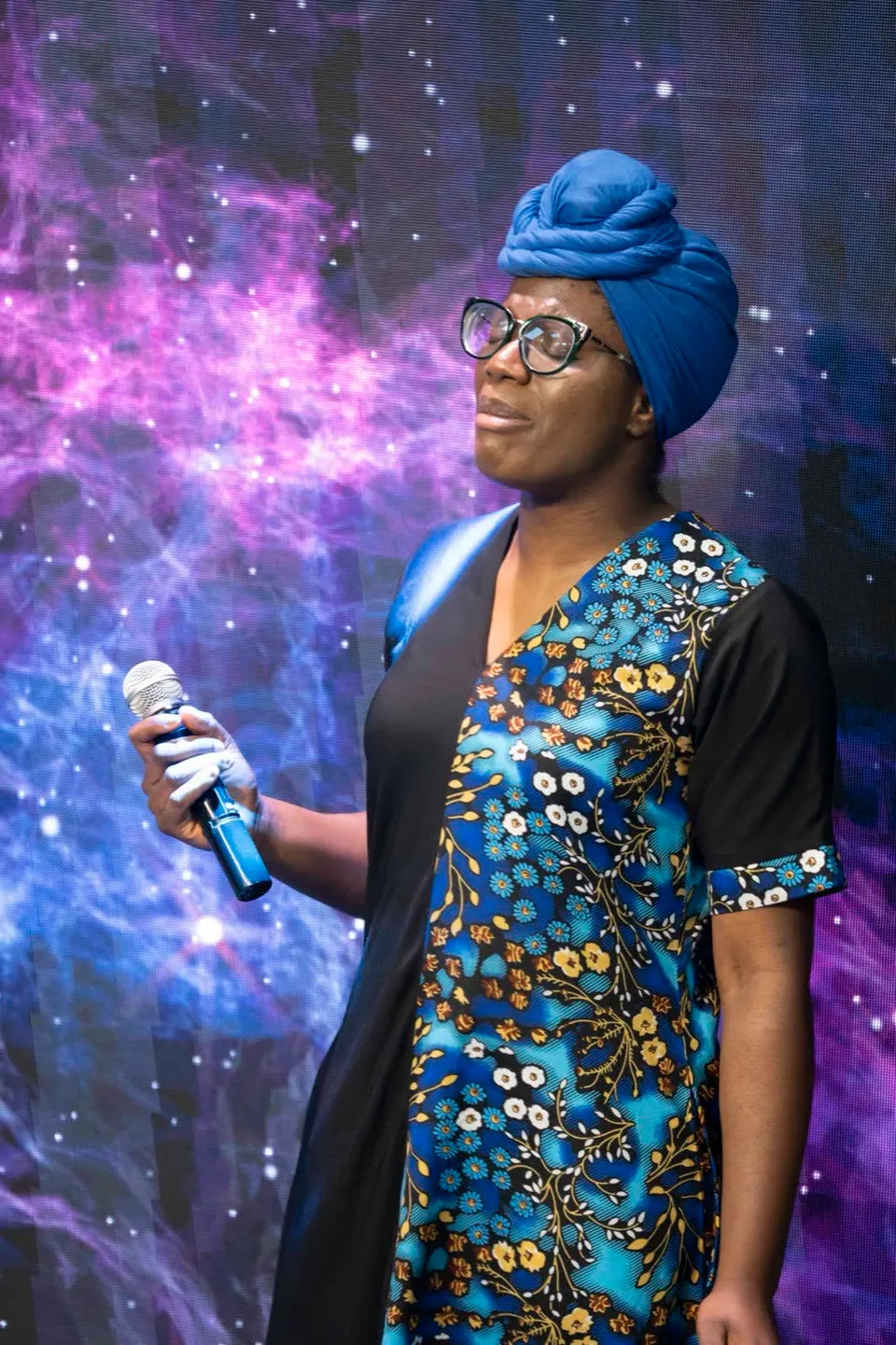 Woman in a blue head wrap and floral dress holding a microphone in worship, eyes closed, at Kingdom Stars Assembly Guelph