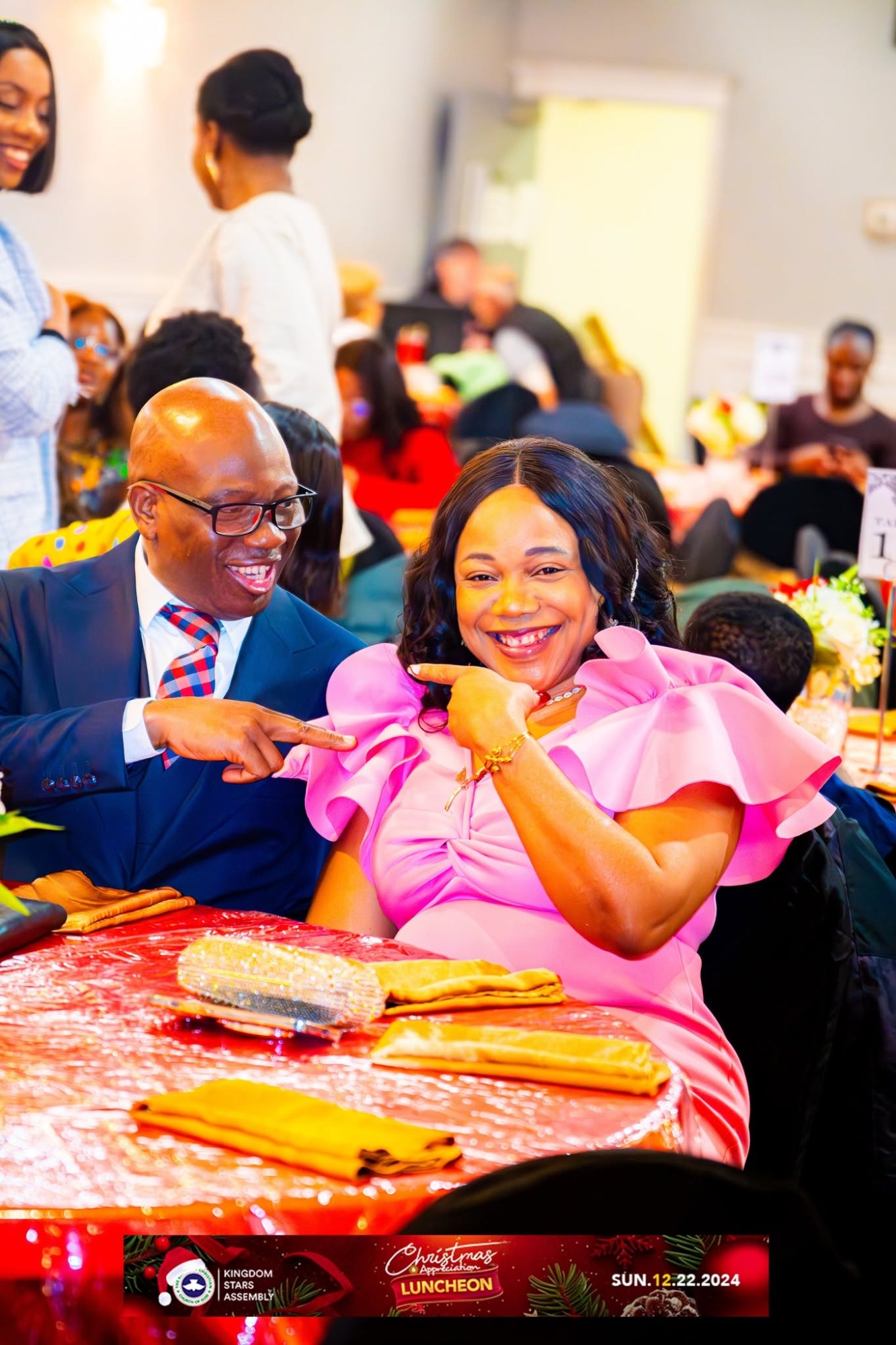 A man in a suit and a woman in a pink ruffled dress laugh and playfully point at each other at the Kingdom Stars Assembly Christmas Appreciation Luncheon, seated at a festive red table.