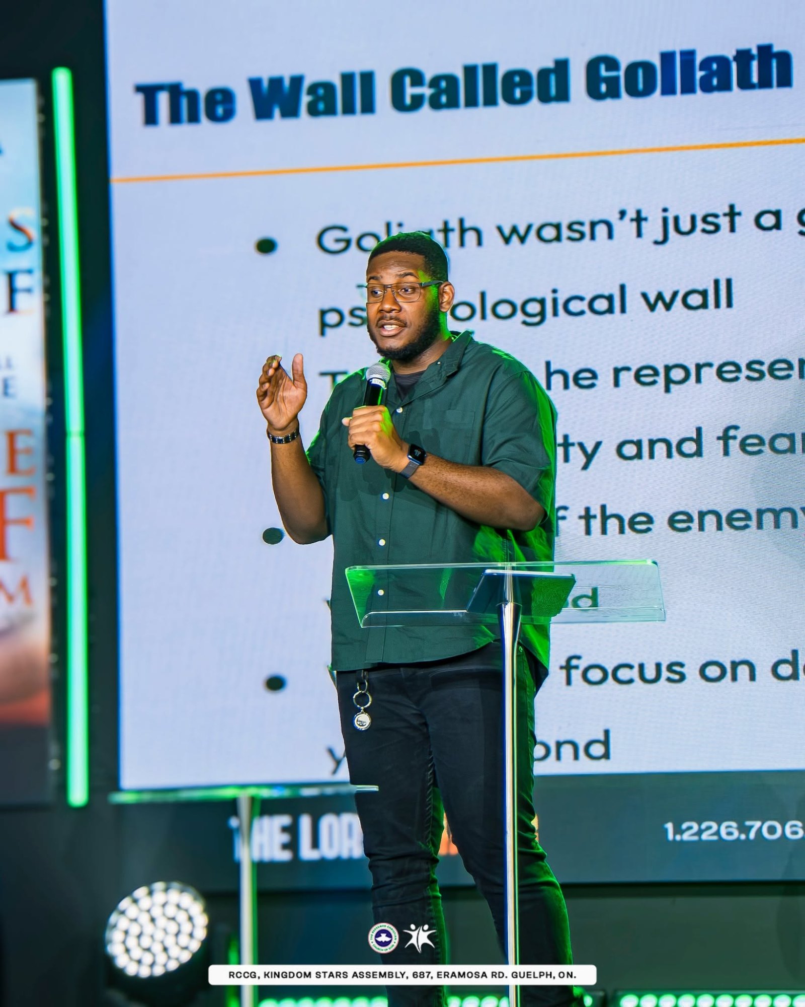 A speaker in a green shirt holds a microphone and gestures while preaching at Kingdom Stars Assembly, with a slide titled "The Wall Called Goliath" on the screen behind him.